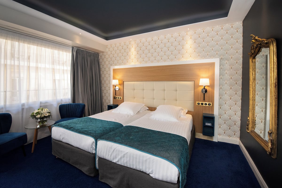 A hotel room with two beds, a sitting area with a chair, a small table with a vase of flowers, and a large mirror on the wall.