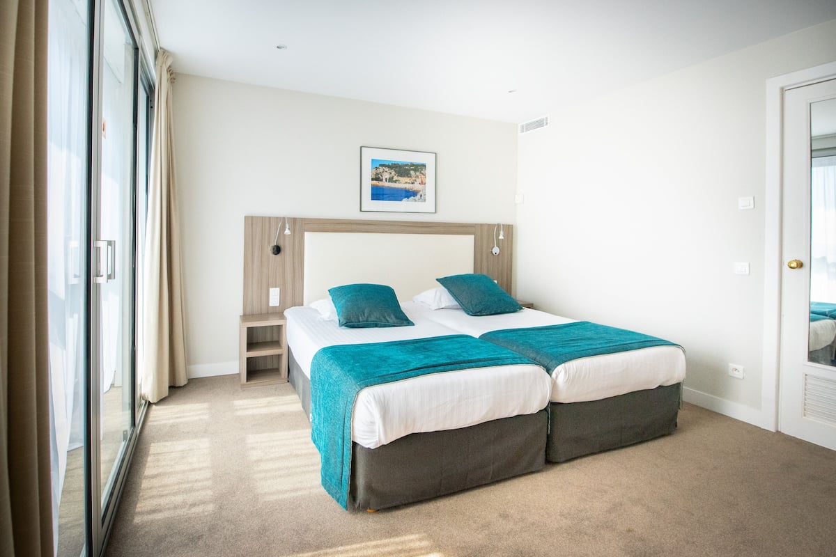 A double bed with a headboard, two pillows, a teal blanket, and a small bedside table.
