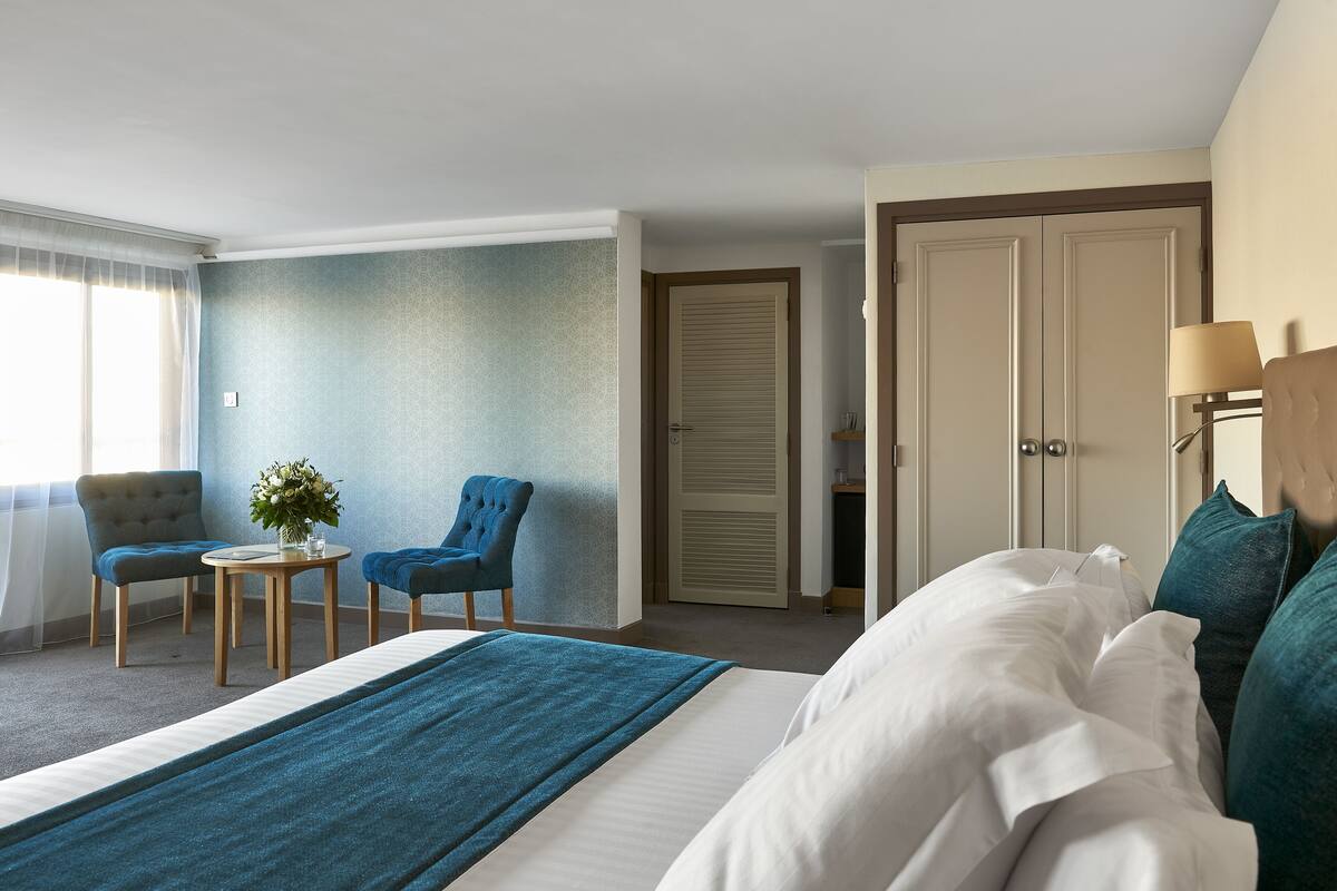 A hotel room with a large bed, two armchairs, a small table with a vase of flowers, a lamp, and a wardrobe.