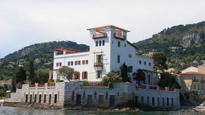 A white building with a tower, located by a body of water, surrounded by greenery and hills. - Splendid Hotel & Spa Nice (Nice)