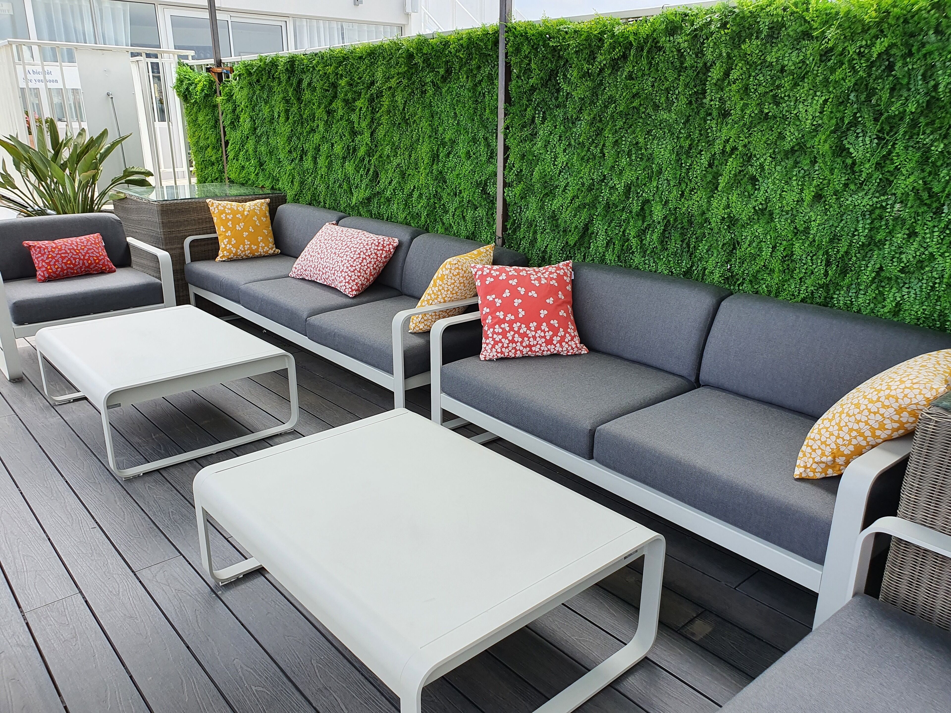 A modern outdoor seating area with grey sofas and white tables, surrounded by a green hedge wall.