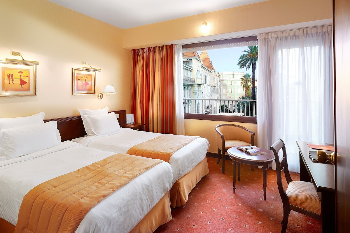 A hotel room with two beds, a small table, a chair, a window with curtains, and artwork on the walls.