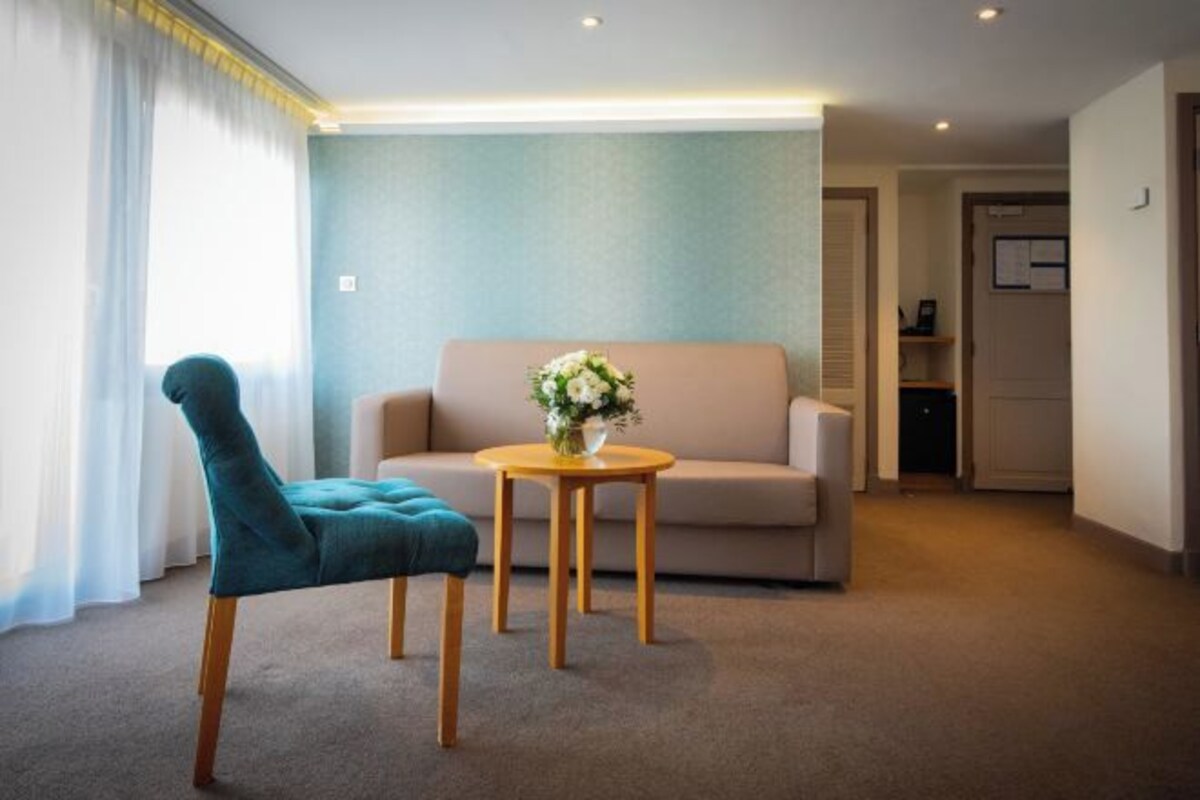 A living room with a teal chair, a beige sofa, and a small round table with a vase of flowers.