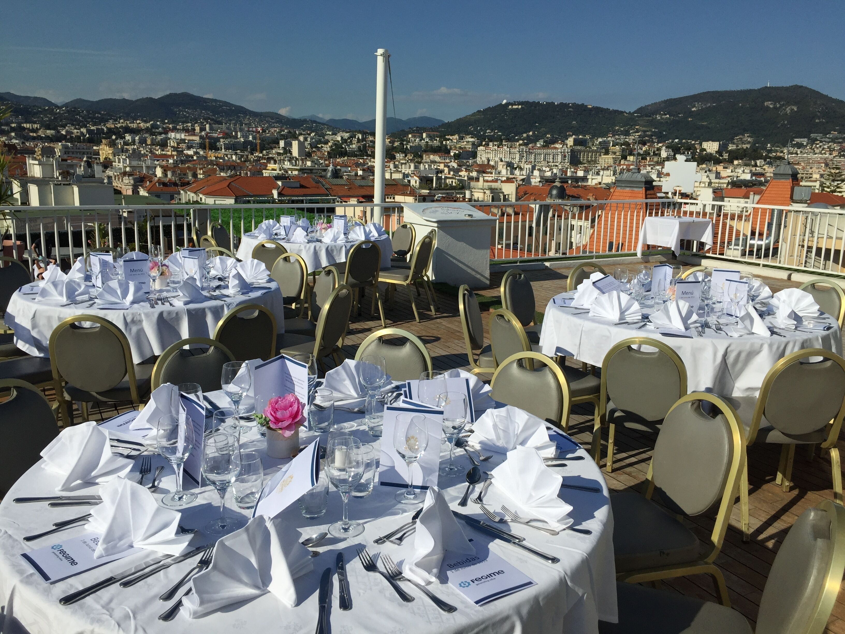 Un restaurante en la azotea con mesas redondas preparadas para comer, con vistas a un paisaje urbano y montañas al fondo.