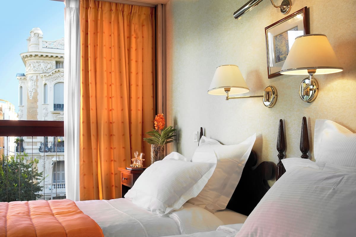 A hotel room with a bed, a nightstand, two wall-mounted lamps, a vase with flowers, and a view of a building outside.