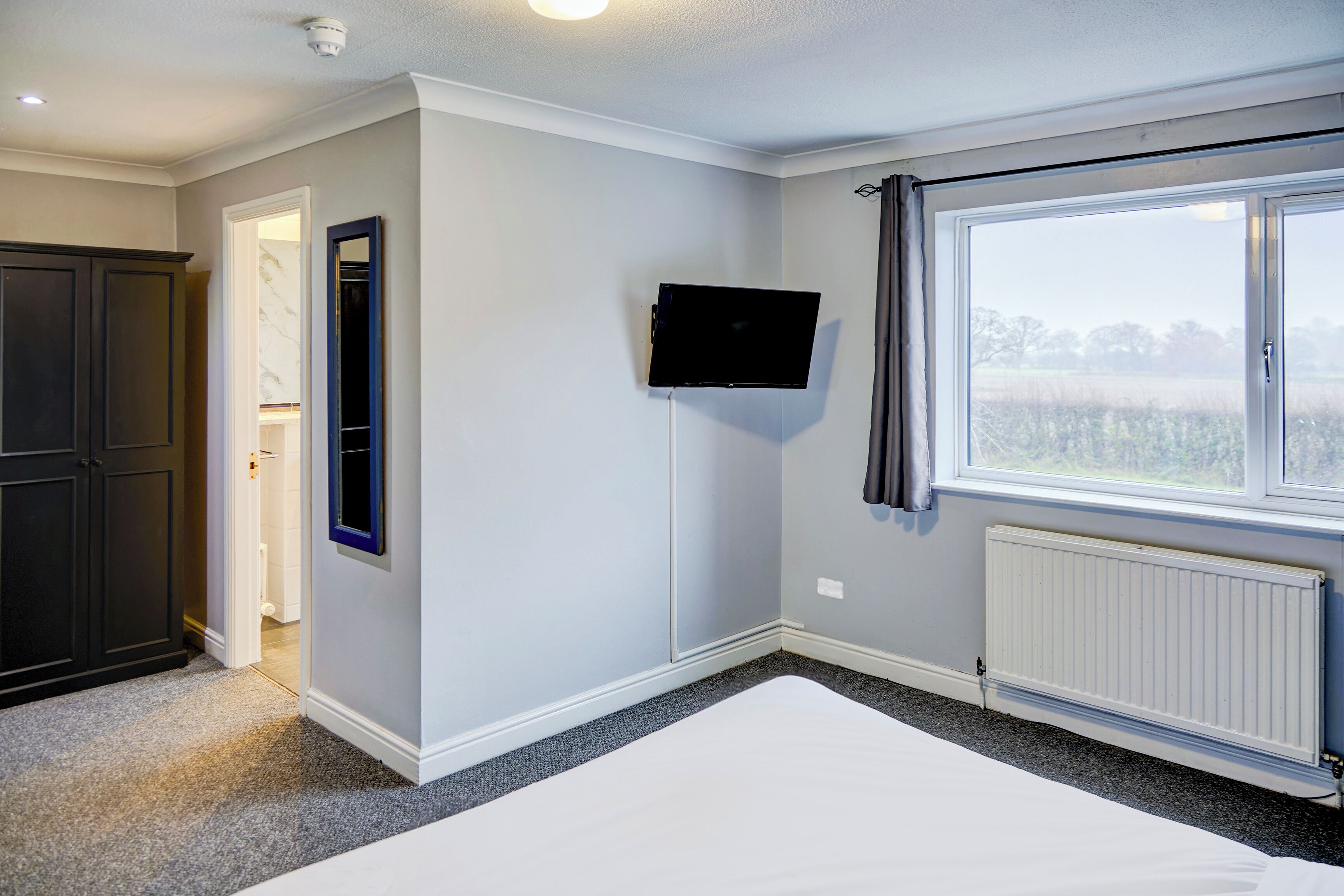 Un dormitorio con cama, televisor montado en la pared, ventana con cortinas y un radiador.