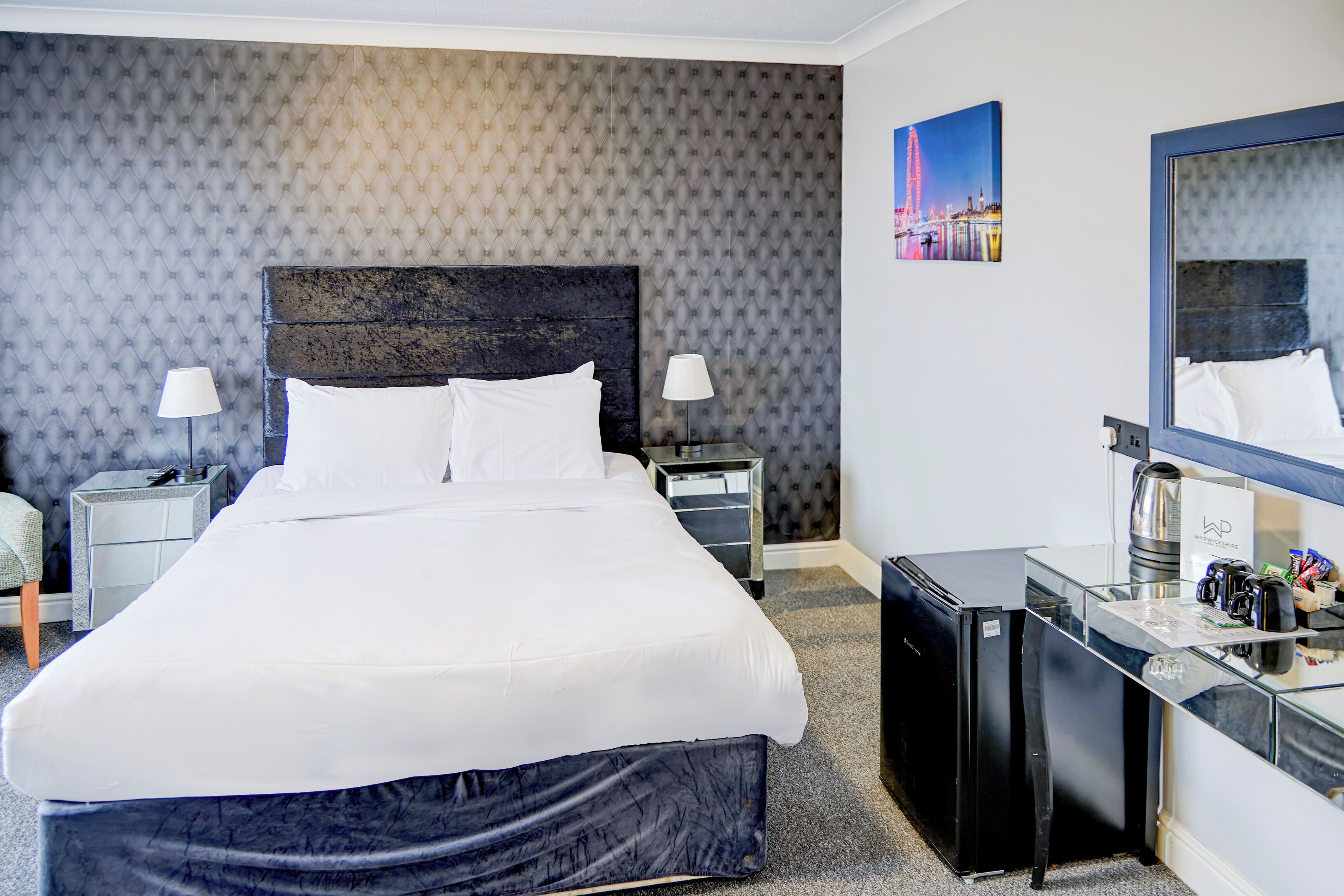 A hotel room with a large bed, a desk, a refrigerator, and a mirror.