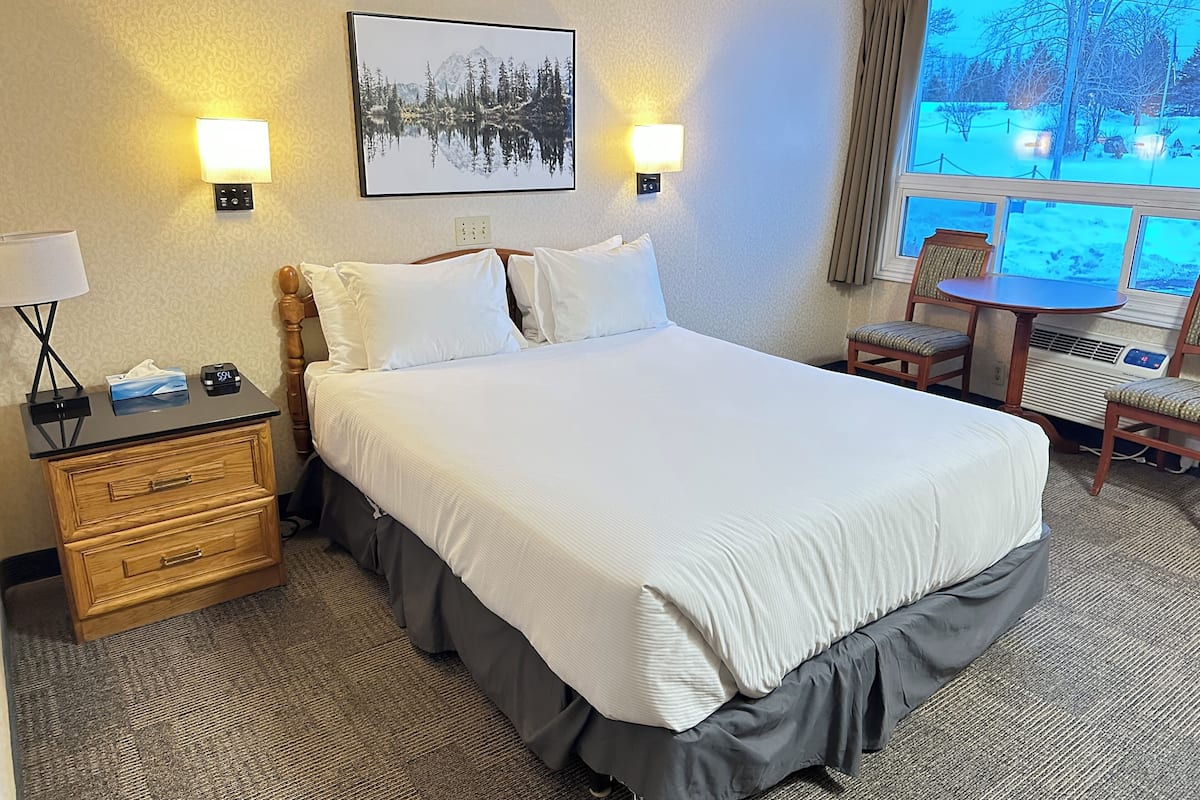Habitación de hotel con una cama grande, lámparas de noche, un escritorio con lámpara, una silla y una ventana con vistas a un paisaje nevado.