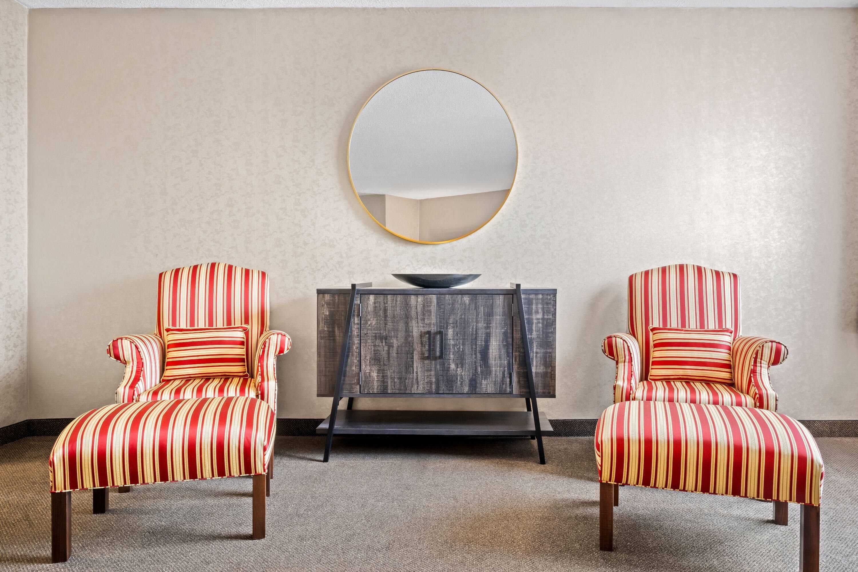 A room with two striped armchairs, a round mirror, and a wooden sideboard.