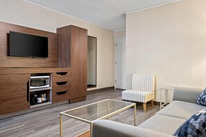 A modern living room with a flat-screen TV, built-in microwave, wooden cabinet, glass coffee table, striped armchair, and a sofa. - Travelodge by Wyndham Amherst (West Amherst)