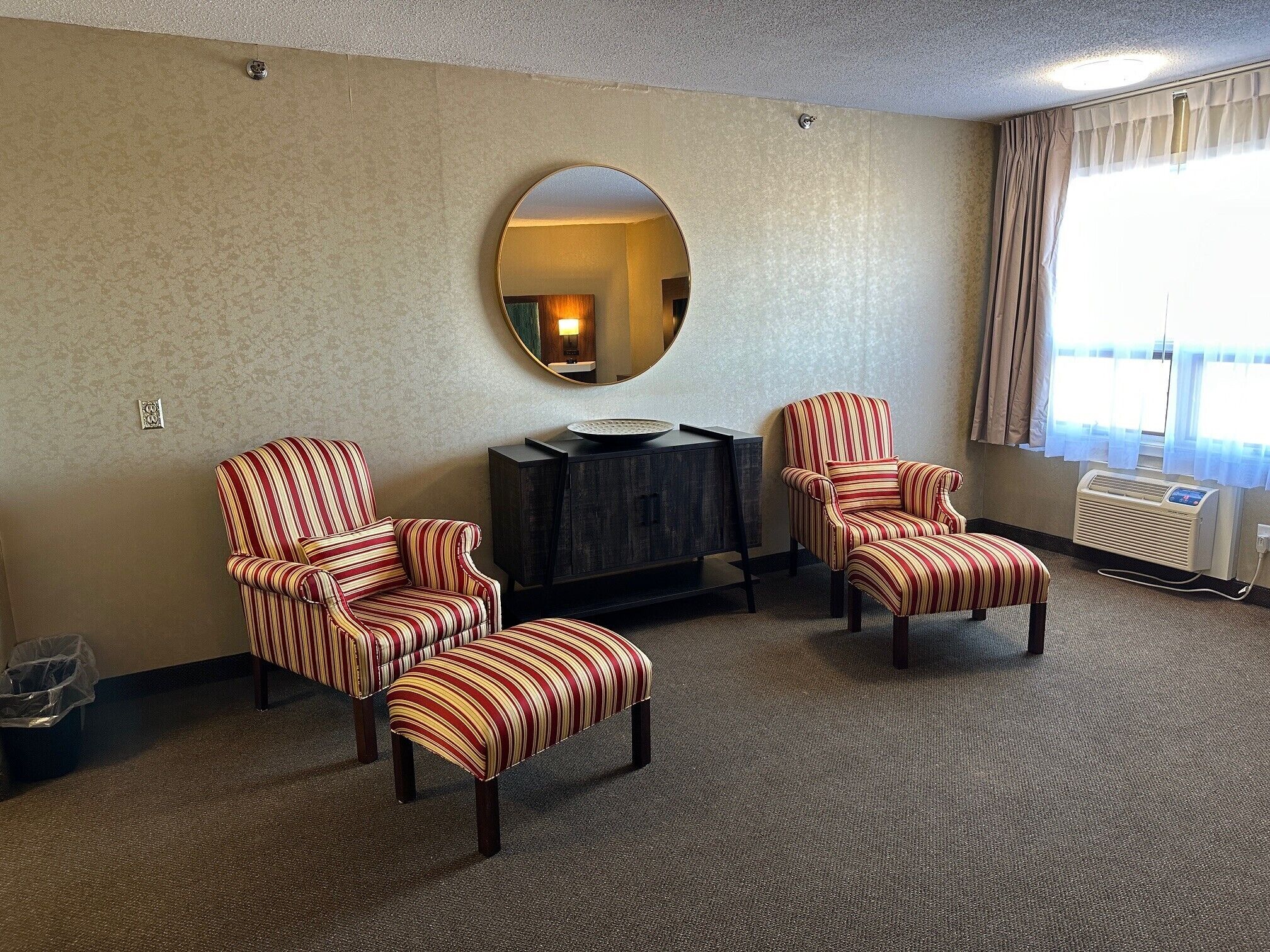 A hotel room with striped armchairs, a round mirror, a dark wooden cabinet, and a radiator.