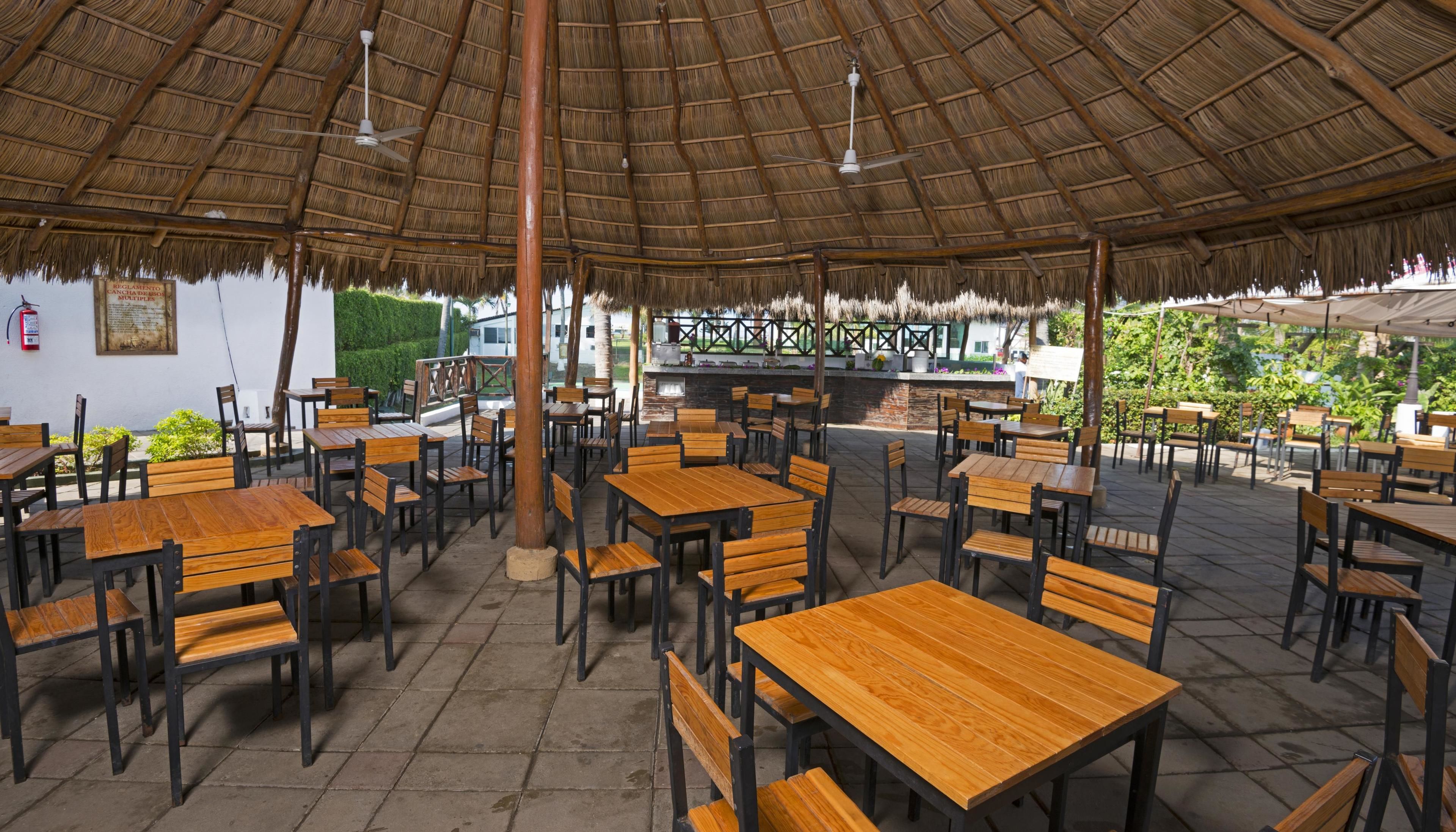 An outdoor restaurant with wooden tables and chairs, a thatched roof, and a bar area.