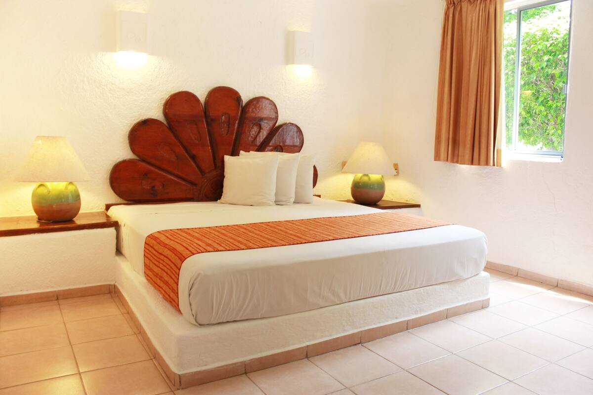 A neatly arranged bedroom with a large bed, two bedside lamps, a decorative wooden headboard, and a window with curtains.