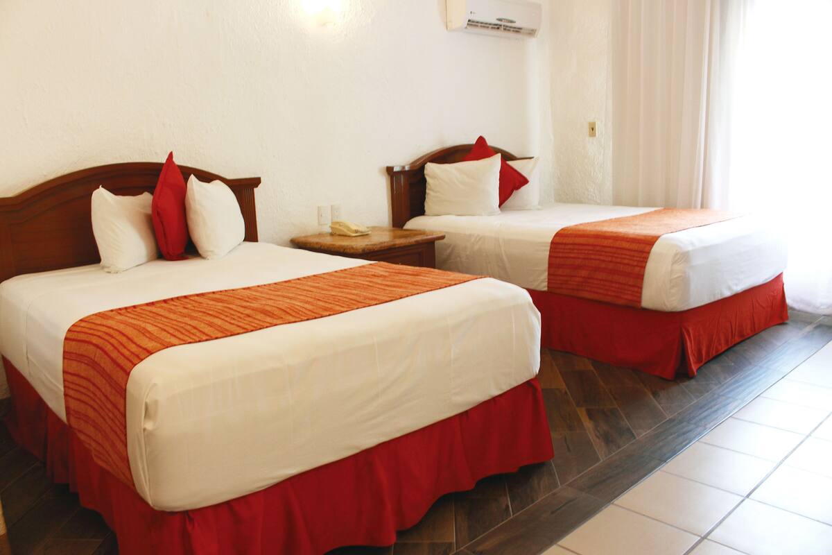 Two beds with red and white bedding, a wooden headboard, a nightstand with a phone, and a white wall with an air conditioning unit.