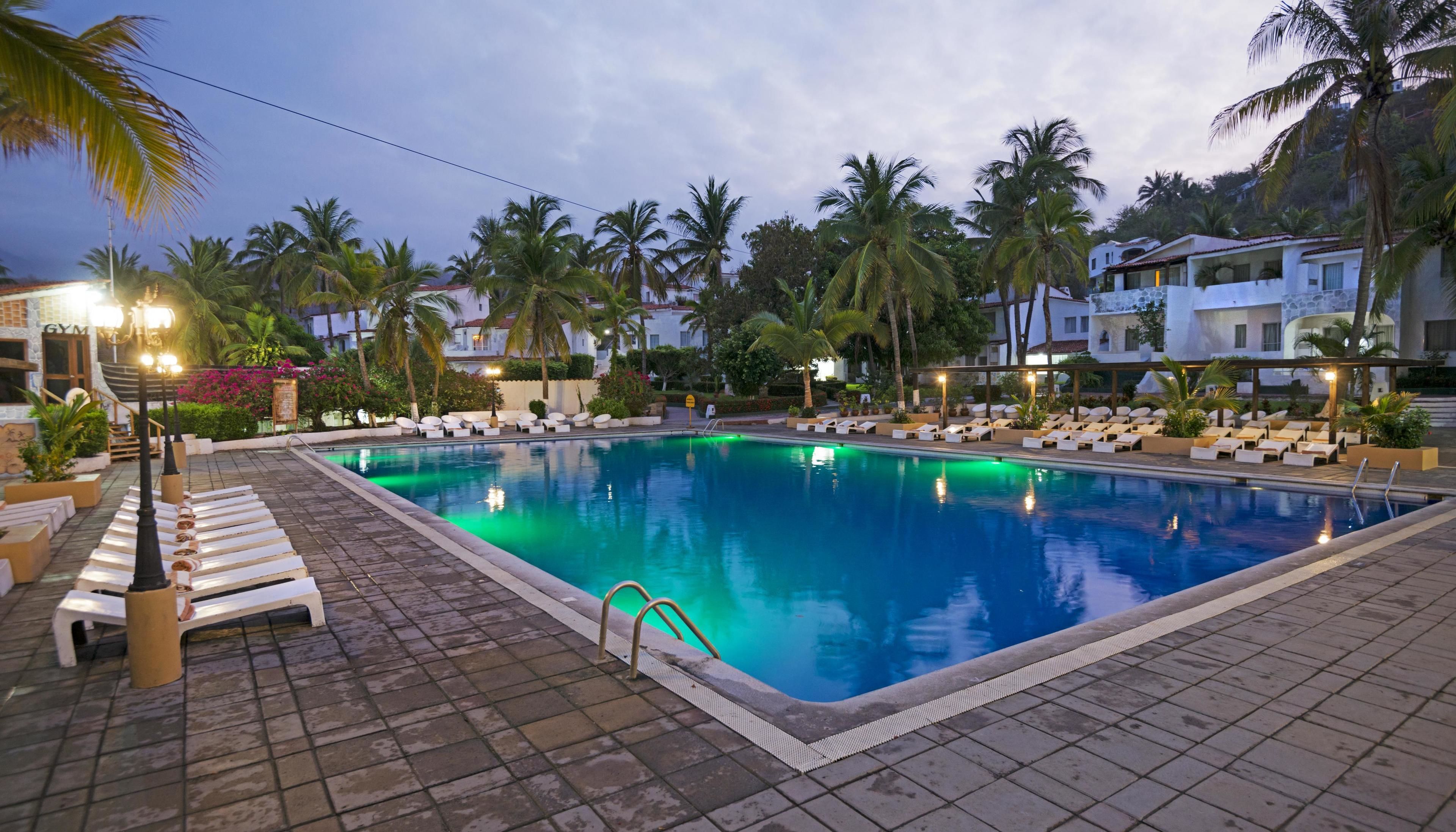 A swimming pool with lounge chairs and palm trees, surrounded by buildings.