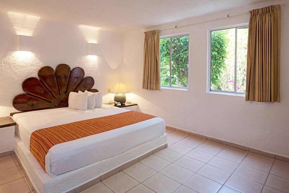 A hotel room with a large bed, two bedside tables with lamps, a decorative headboard, and two windows with curtains.