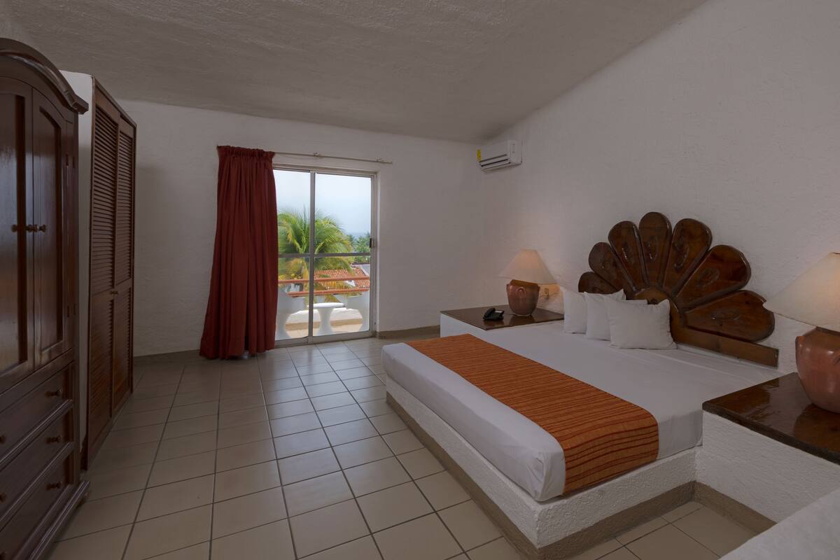 A hotel room with a bed, a nightstand, a wooden headboard, a wardrobe, a sliding glass door to a balcony, and a ceiling fan.