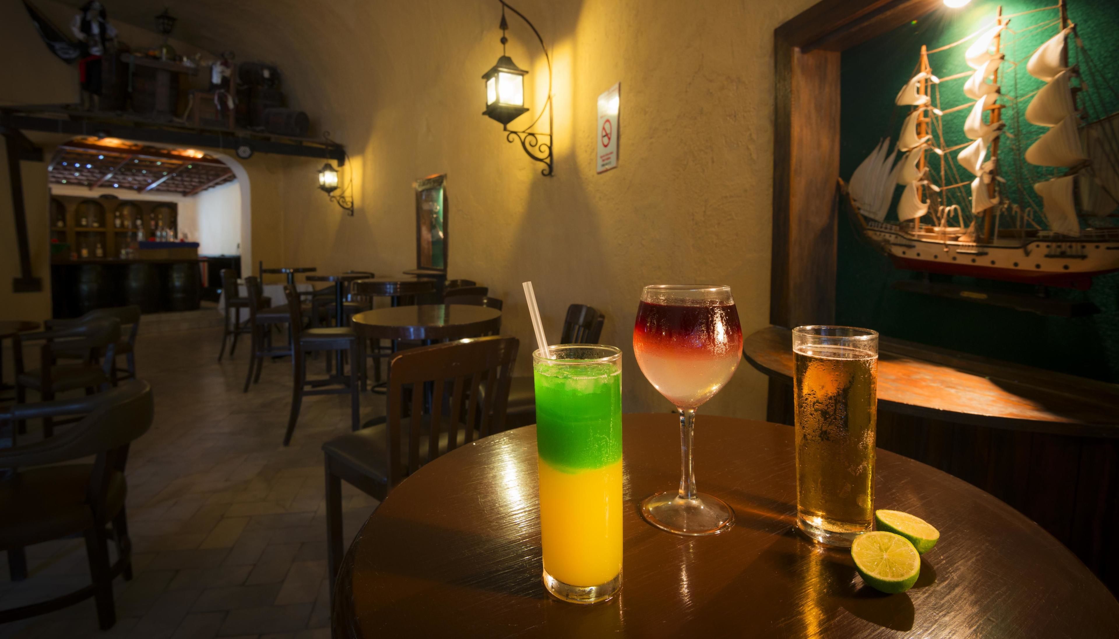 Zona de bar con mesas de madera, una bebida verde, una copa de vino y una jarra de cerveza.