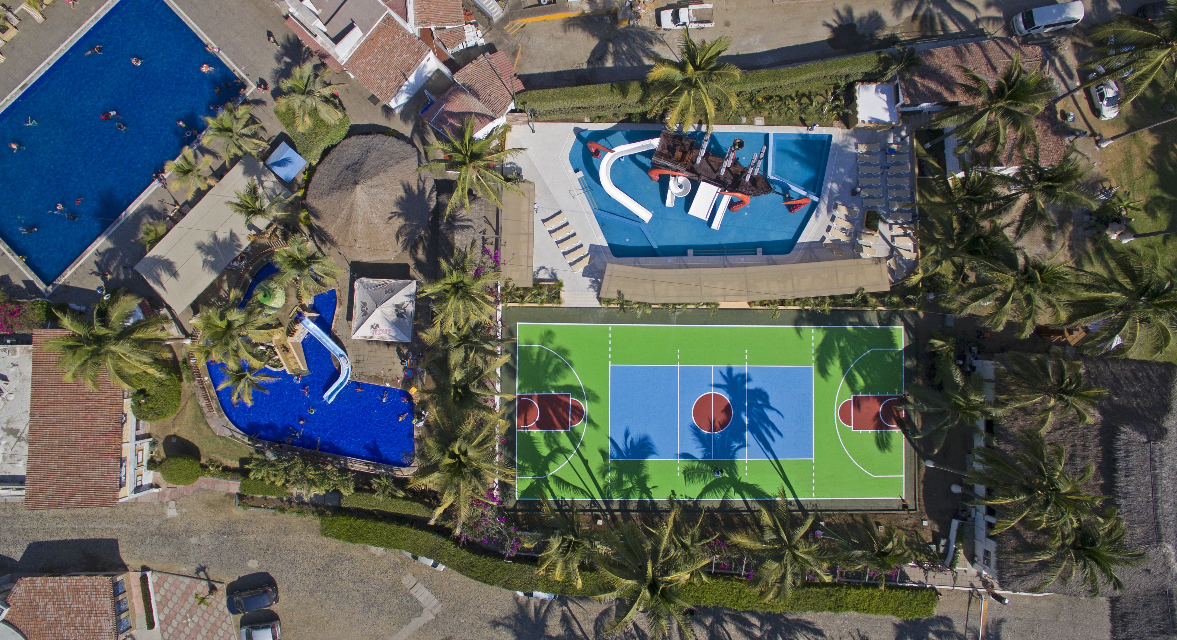 An aerial view of a resort with a swimming pool, a basketball court, and palm trees.