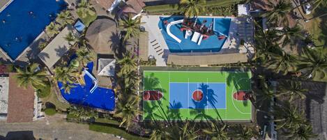 An aerial view of a resort with a swimming pool, a basketball court, and palm trees.