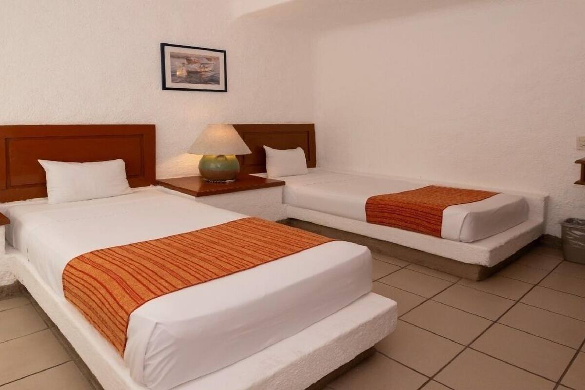 A hotel room with two beds, a nightstand with a lamp, a framed picture on the wall, and a trash bin.