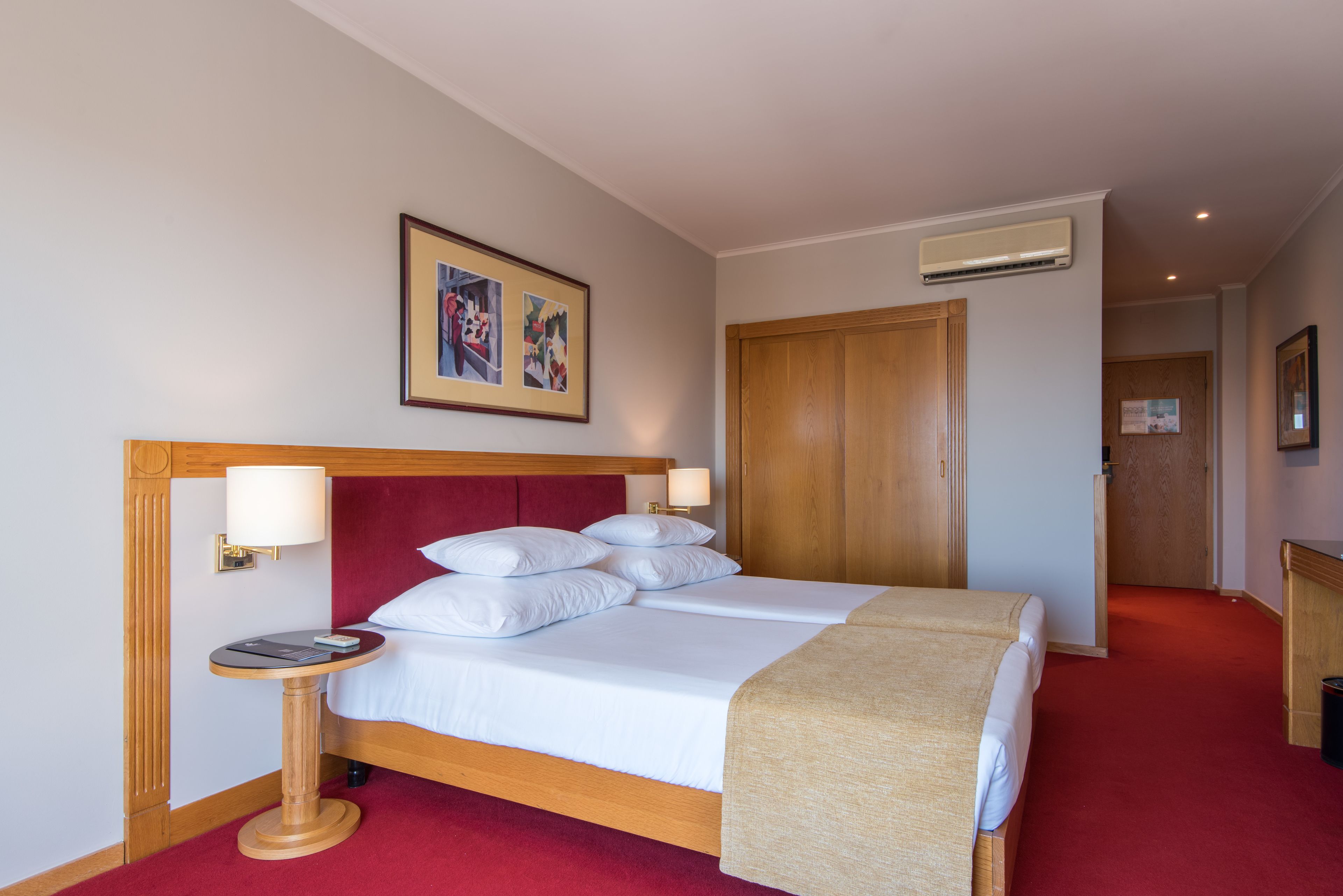 Een hotelkamer met een groot bed, een nachtkastje met een lamp, een ingelijst schilderij aan de muur en een houten deur.