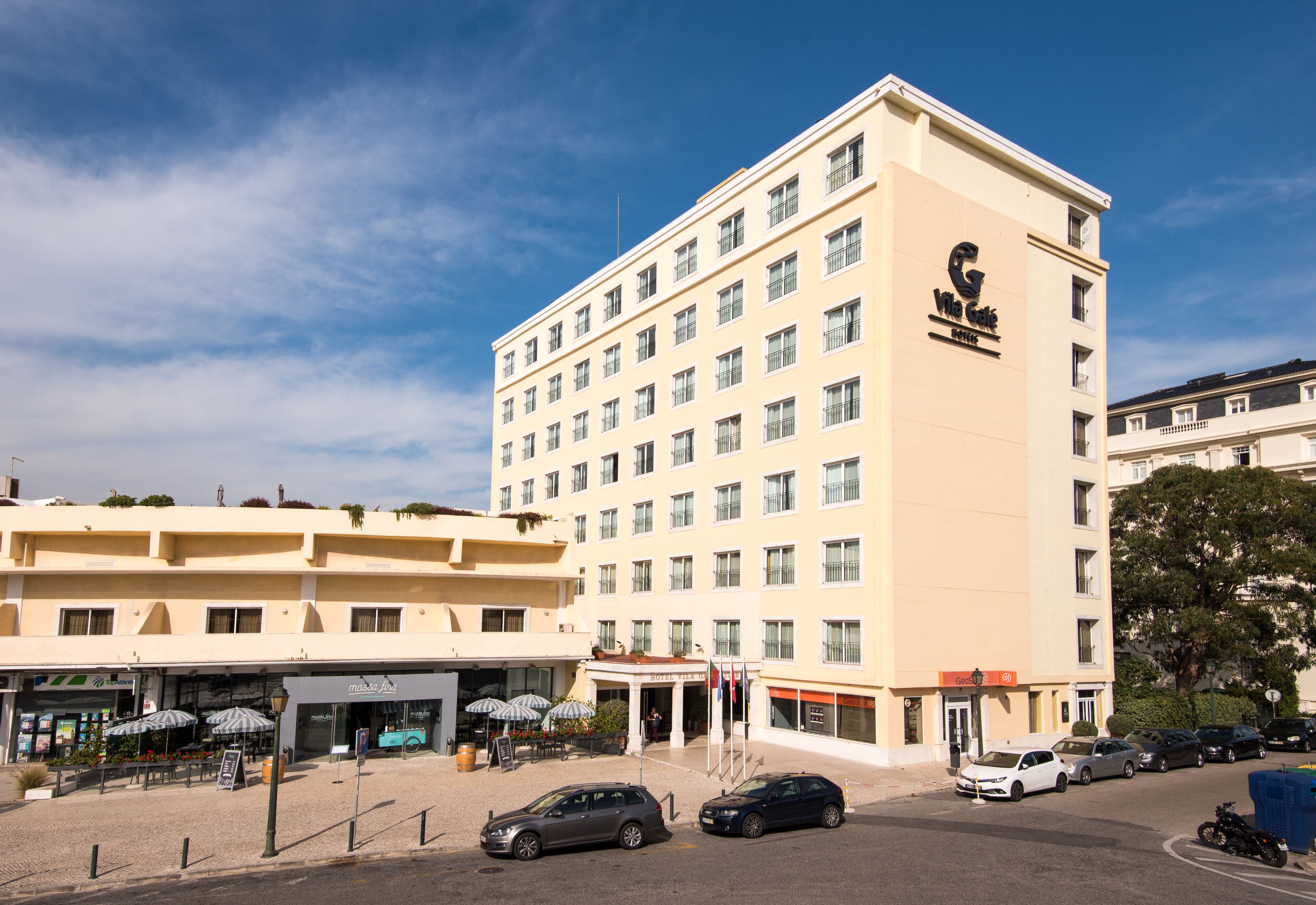 A multi-story hotel with a prominent logo on the facade, a parking area with several cars, and outdoor seating with umbrellas.