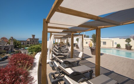 A rooftop pool area with sun loungers and umbrellas, overlooking a cityscape and the sea.