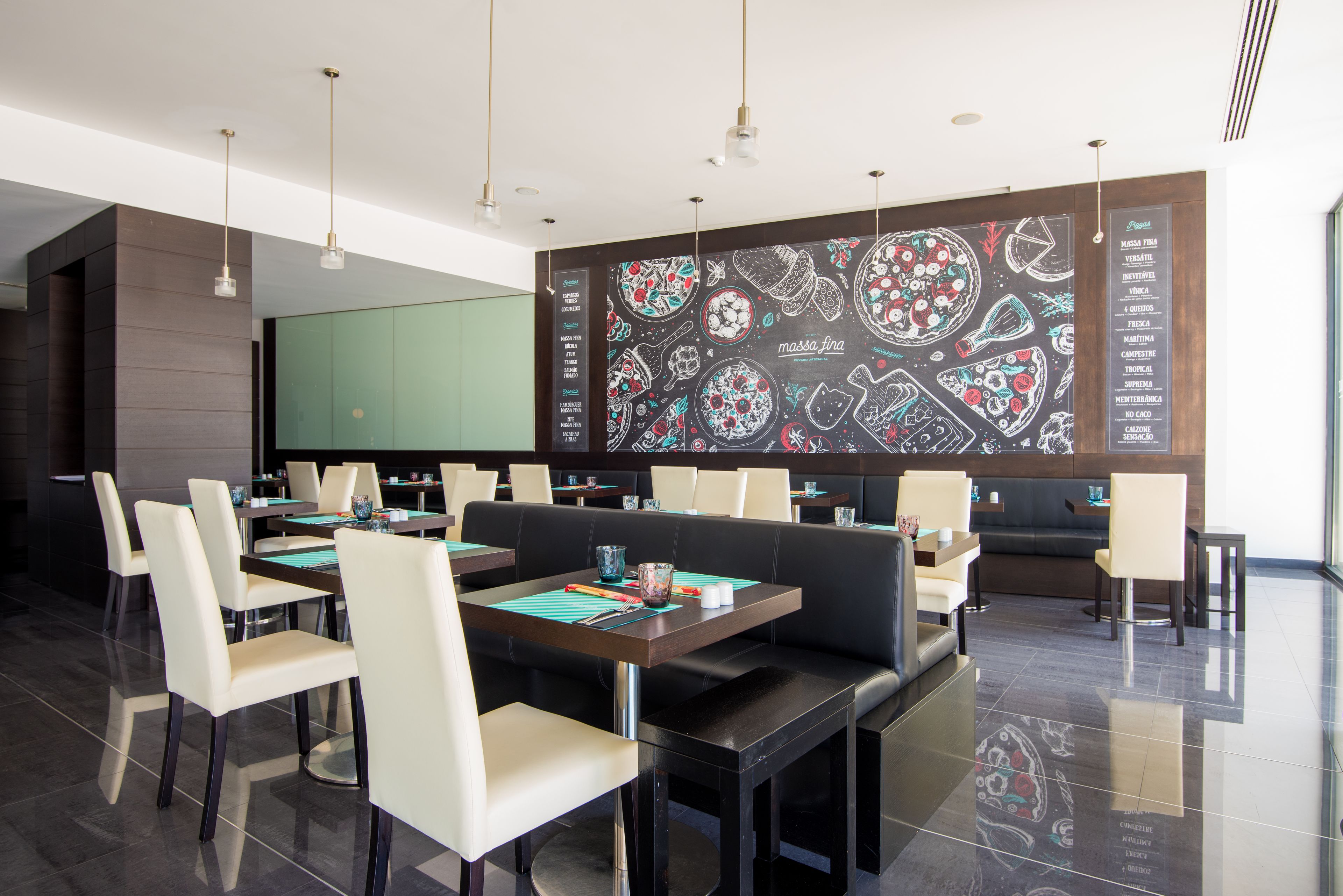 Un restaurante moderno con techo alto, iluminación colgante y una pared con menú en pizarra. Las mesas están dispuestas con sillas, y hay una zona de estar con sofá.