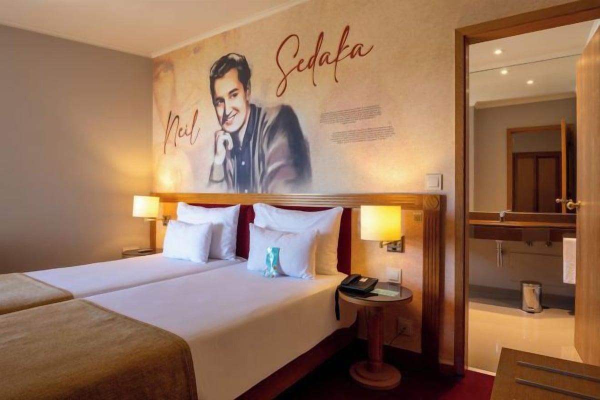 A hotel room with a bed, two bedside lamps, a nightstand, a mirror, and a wall with a portrait.