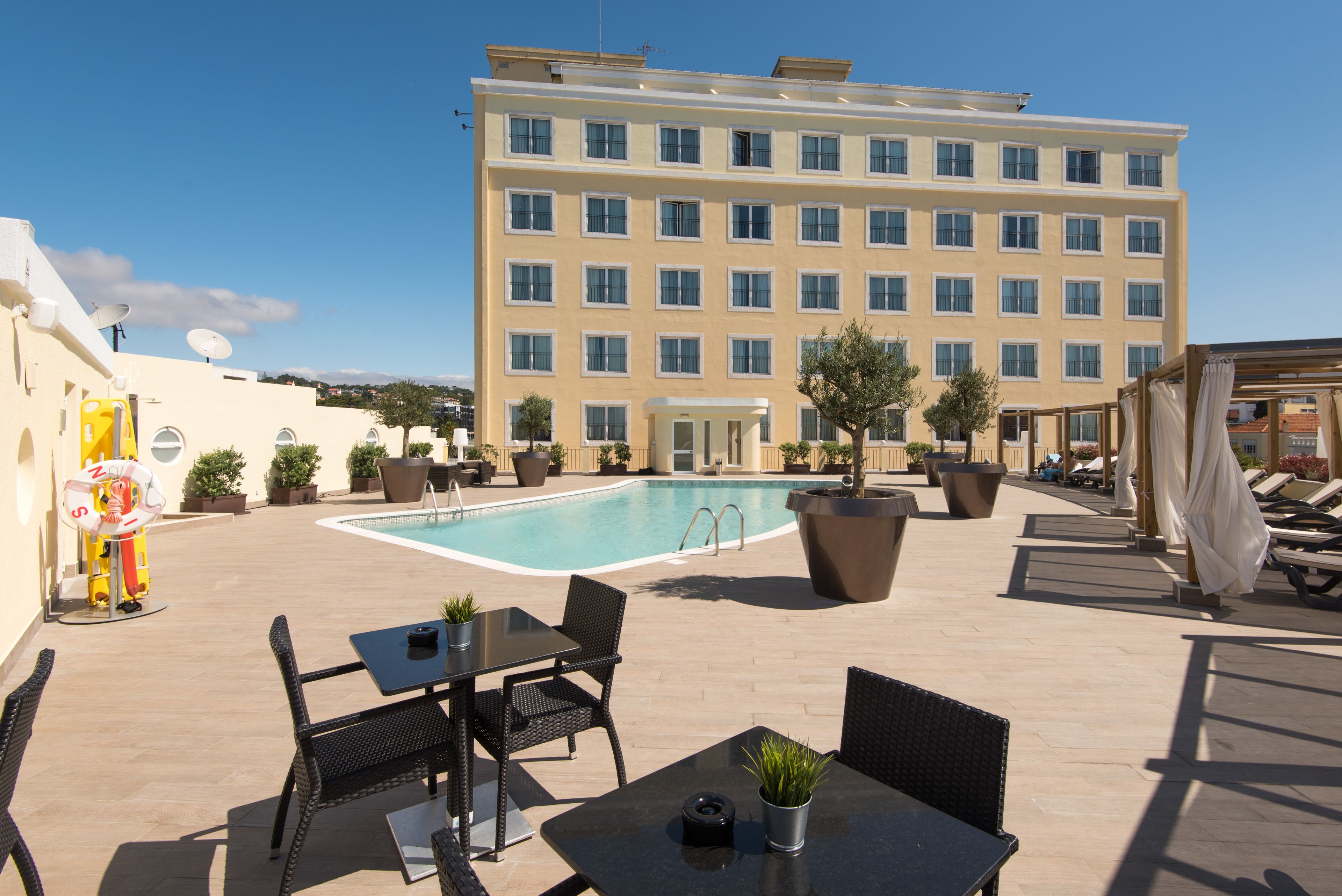 A rooftop pool area with a building in the background, outdoor seating, and potted plants.