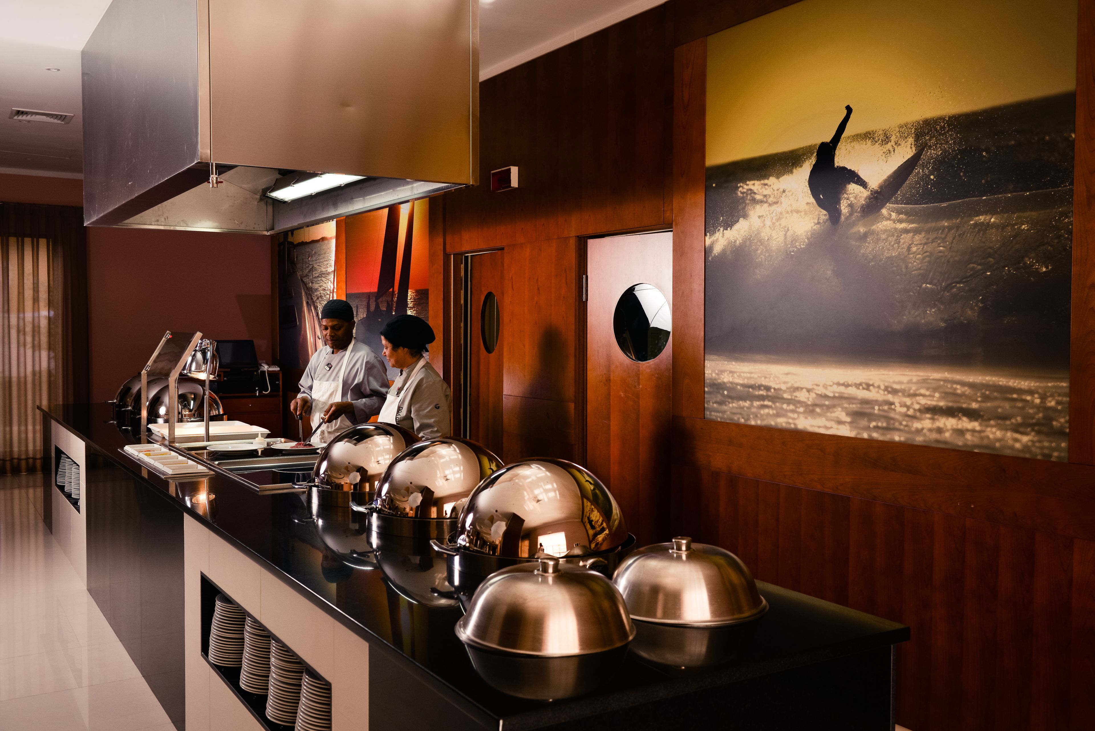 Un buffet avec plusieurs points de service, un chef qui prépare les plats et une grande photo encadrée représentant un surfeur sur une vague.