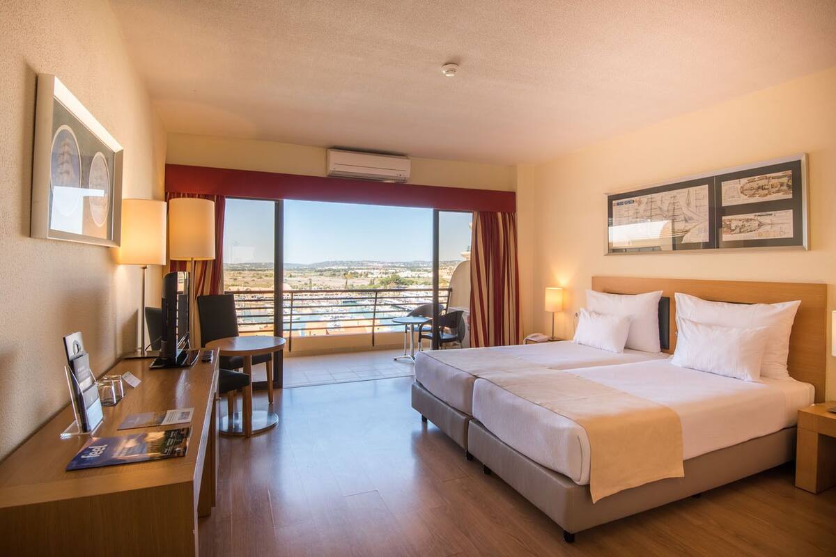 Habitació d'hotel amb un llit gran, una taula de treball amb una cadira, una taula petita i vista al mar a través de grans finestres.