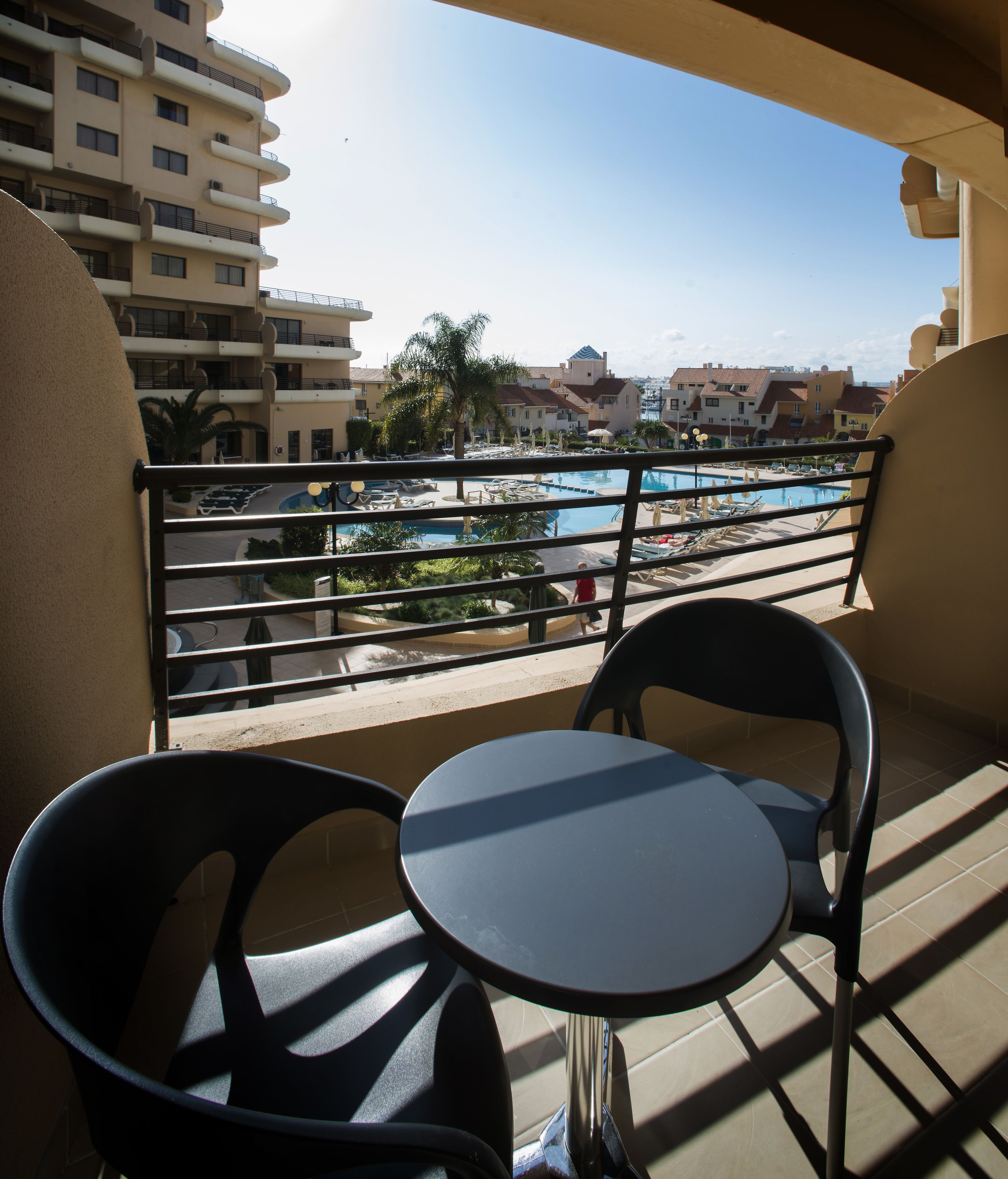A balcony with two chairs and a small table, overlooking a pool area with lounge chairs and umbrellas.