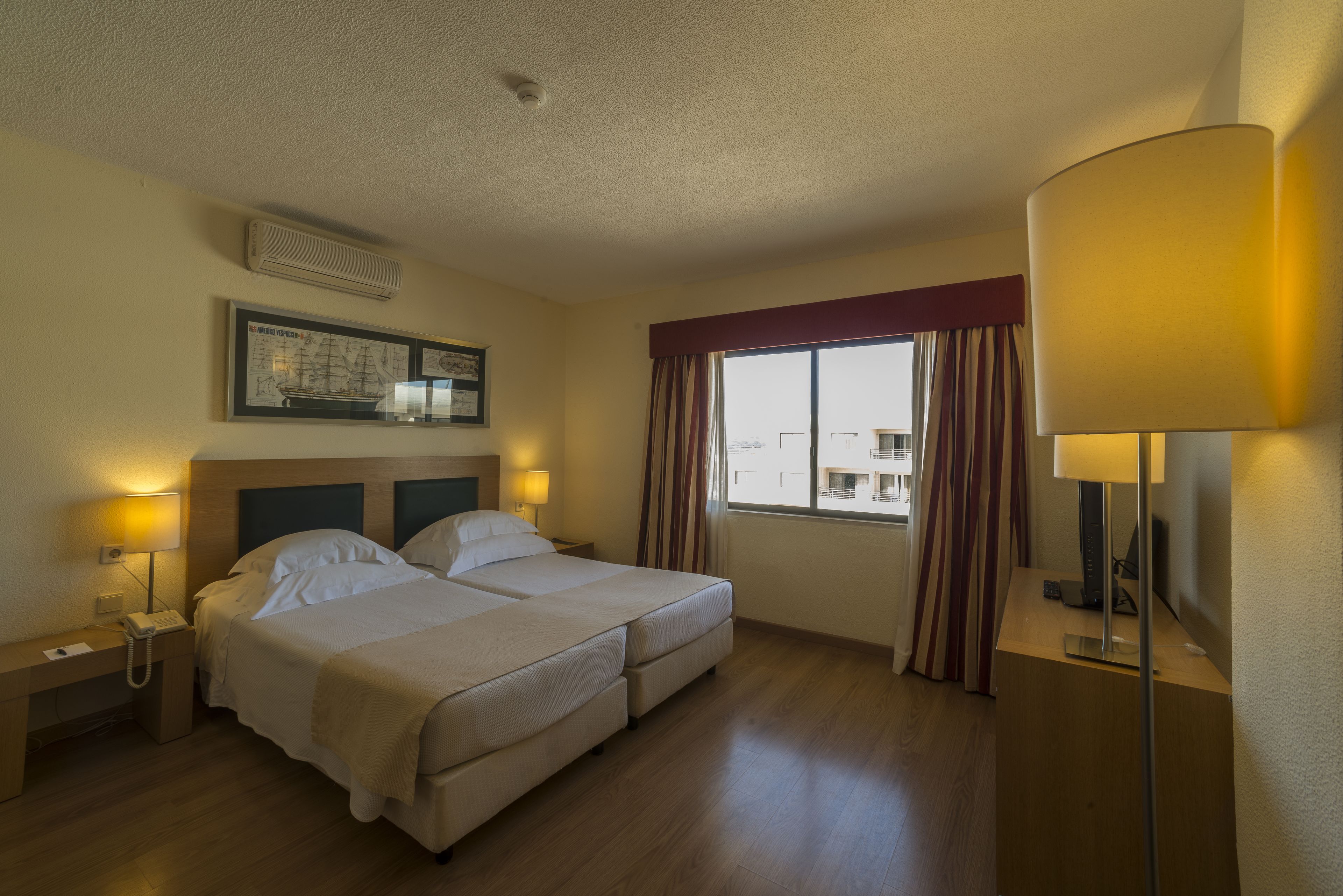 Habitación de hotel con cama doble, lámparas de noche, escritorio con teléfono, televisor de pantalla plana y ventana con cortinas.