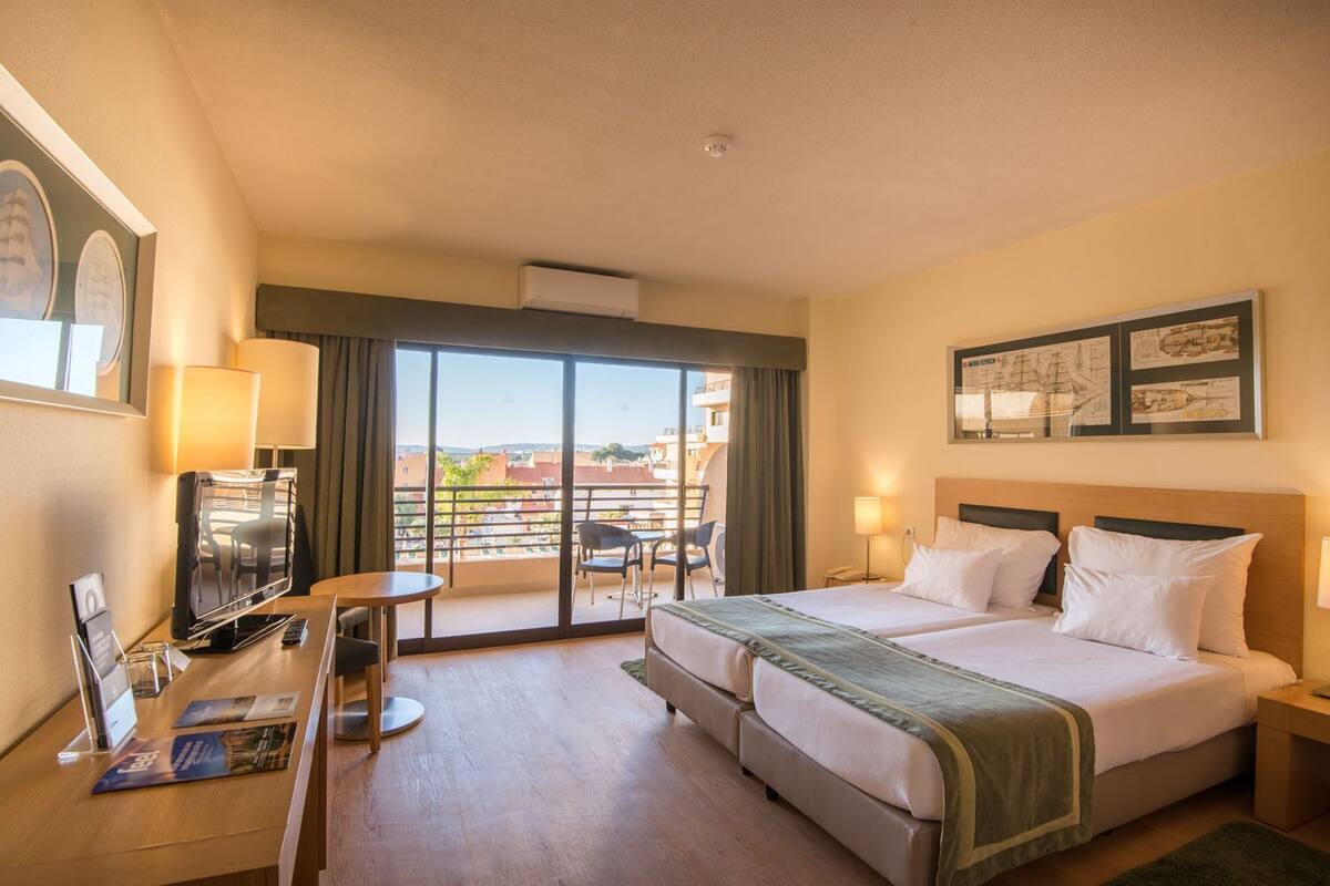 Habitació d'hotel amb un llit, escriptori amb televisió, una cadira, balcó amb taula i cadires, i vistes a edificis.