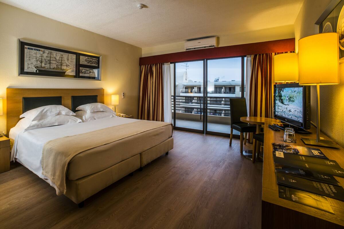Habitació d'hotel amb un llit gran, una taula de treball amb un ordinador, un televisor i un balcó amb vista a la ciutat.