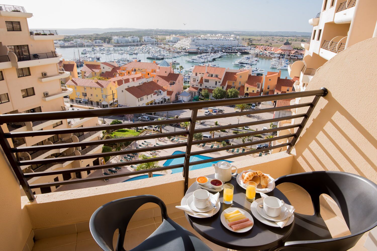 A balcony with a table set for breakfast, overlooking a marina with boats and buildings.