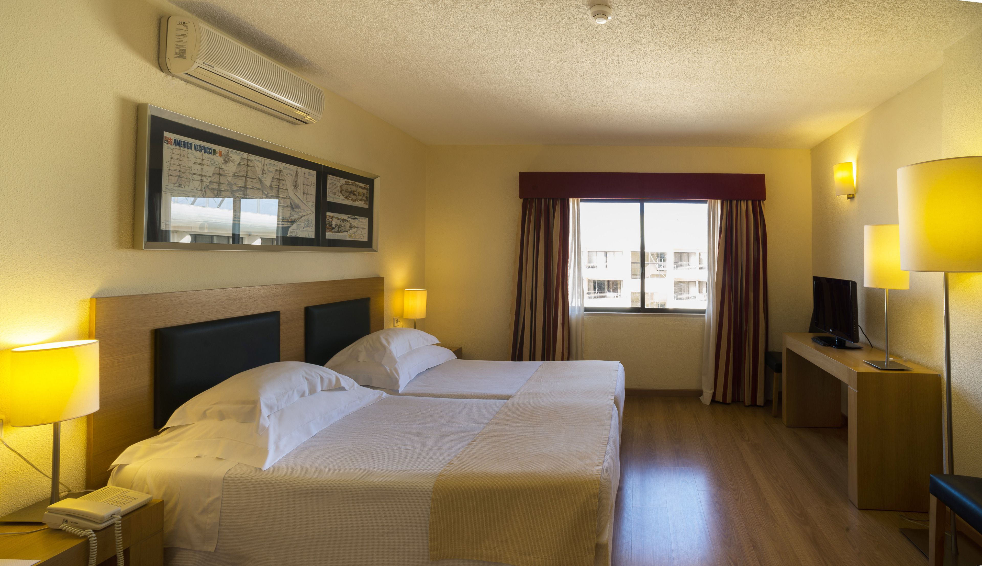 Ett hotellrum med två sängar, ett skrivbord med en dator, en telefon och ett fönster med gardiner.