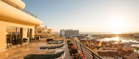 Terraza en la azotea con tumbonas y jardineras de flores, con vistas a un puerto deportivo y edificios.