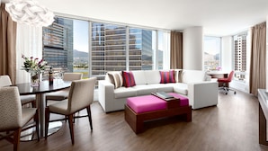 A modern living room with a white sofa, a coffee table, and a dining area with a table and chairs. Large windows offer a view of tall buildings and a body of water. - Auberge Vancouver Hotel (Vancouver)