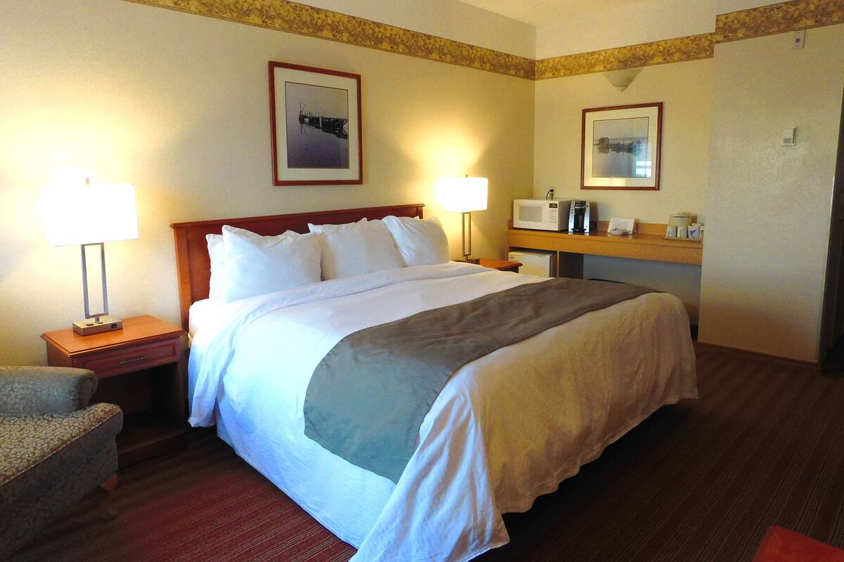 A hotel room with a large bed, two bedside lamps, a nightstand, a chair, a desk with a microwave, and a framed picture on the wall.