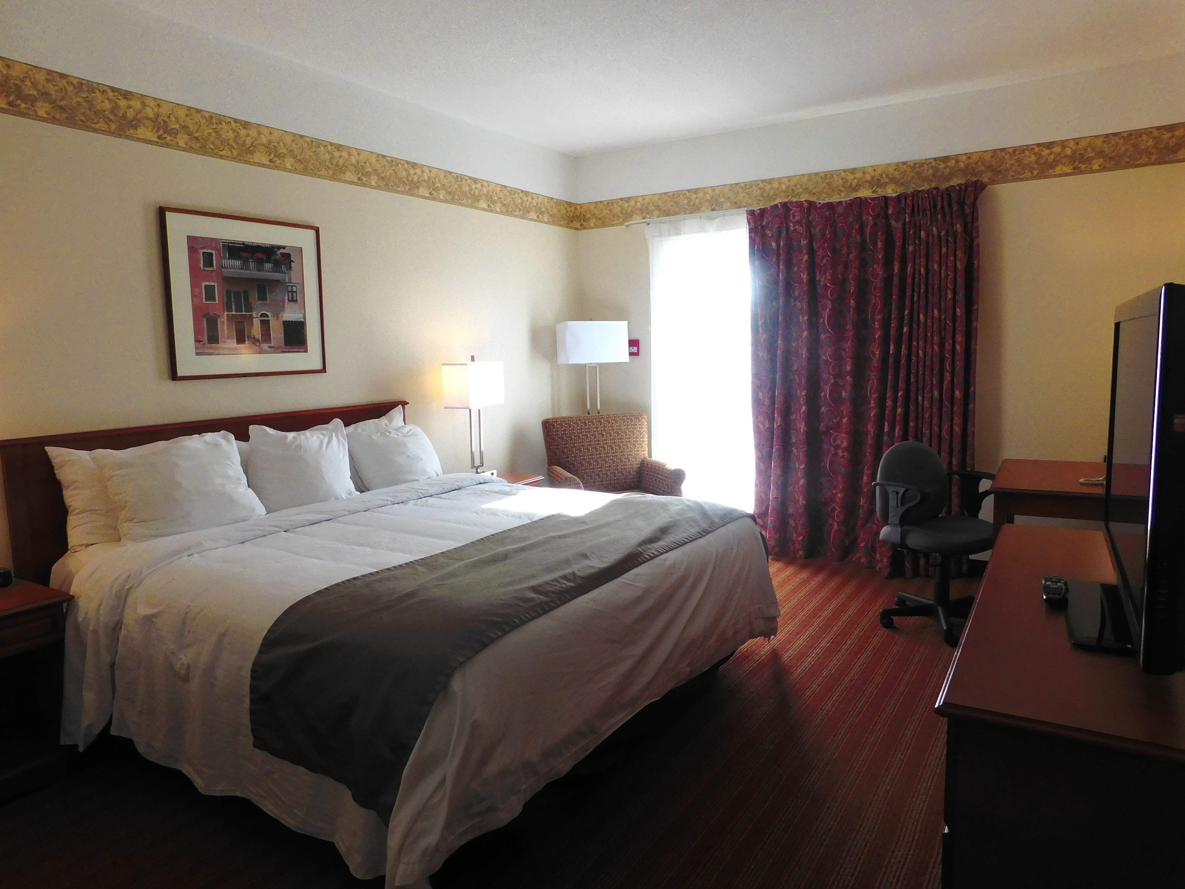 Habitación de hotel con una cama grande, un escritorio con silla, televisor, lámpara y ventana con cortinas.