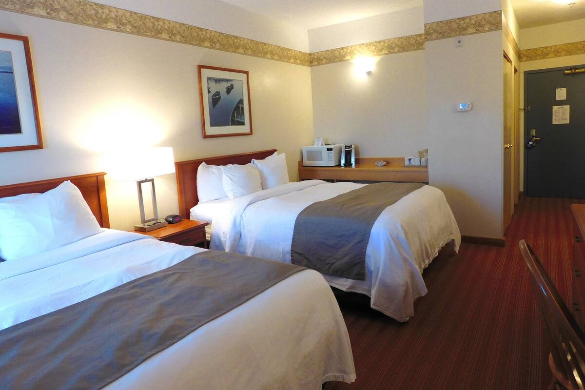 A hotel room with two beds, a bedside table with a lamp, a microwave, and a clock on the wall.