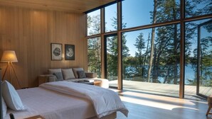 Une chambre à coucher moderne avec un grand lit, une table de chevet, une lampe et une vue sur une forêt, visible à travers de grandes fenêtres.