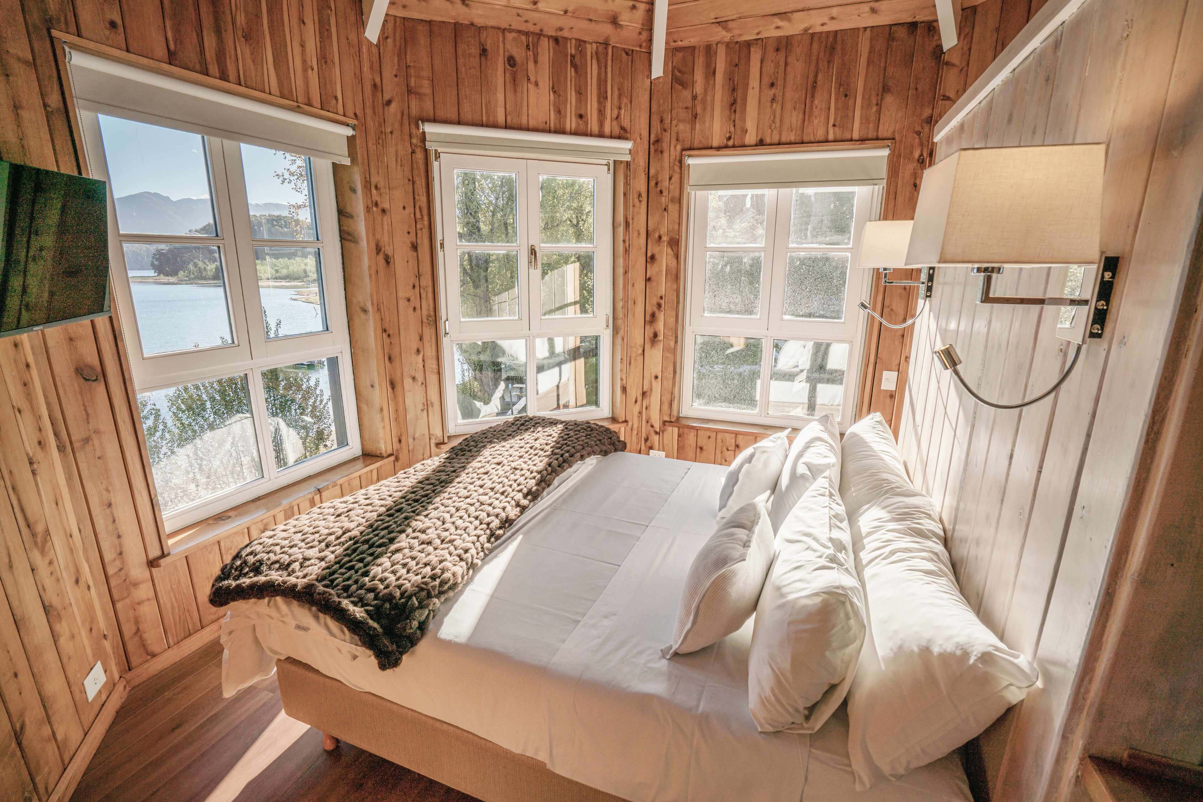 A wooden-paneled bedroom with a bed, two windows, and a wall-mounted lamp.