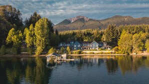 Un chalet au bord du lac, composé de plusieurs bâtiments, avec un quai et des montagnes boisées en arrière-plan.