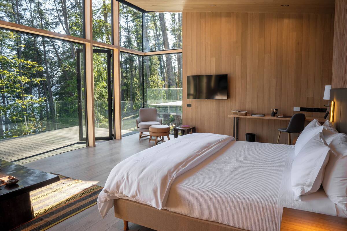 Ein modernes Schlafzimmer mit einem großen Bett, einem Schreibtisch und einem Stuhl. Ein Fernseher ist an der Wand befestigt und ein Fenster bietet Blick auf den Wald.