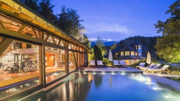 Un bâtiment moderne à la façade vitrée, un espace piscine avec des chaises longues, et une grande maison en arrière-plan.