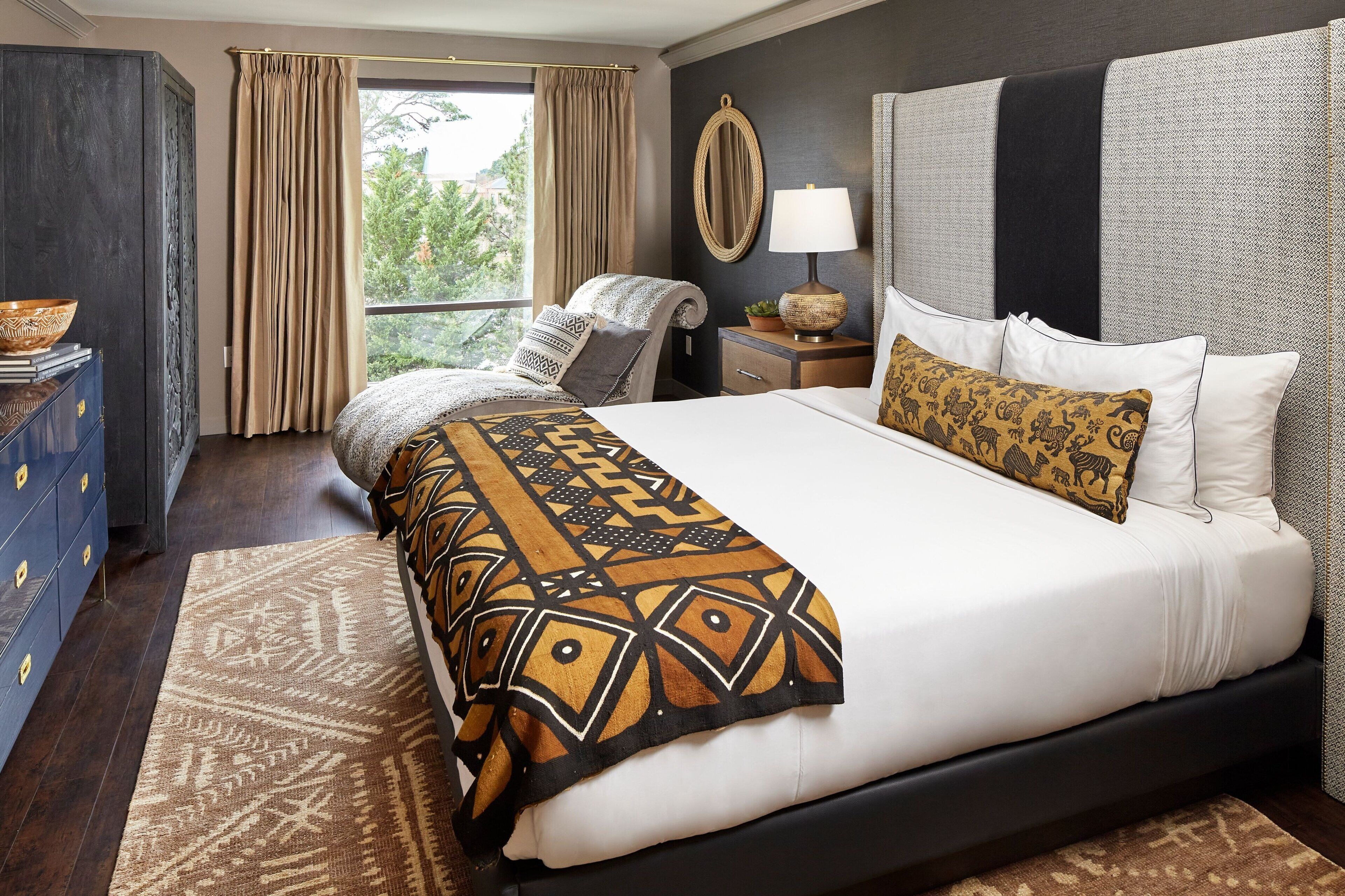 A hotel room with a large bed, a bedside table with a lamp, a dresser, and a view of greenery outside.